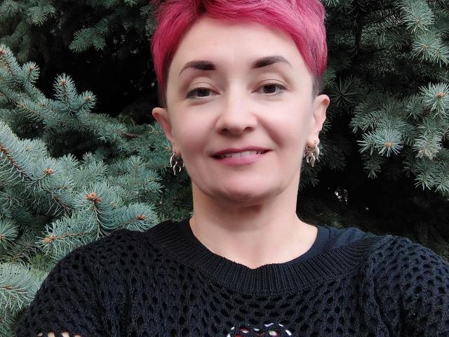 ChristinaRainforce with short pink hair, wearing a black patterned sweater, smiling, earrings, and a cute face, standing in front of a green pine tree.