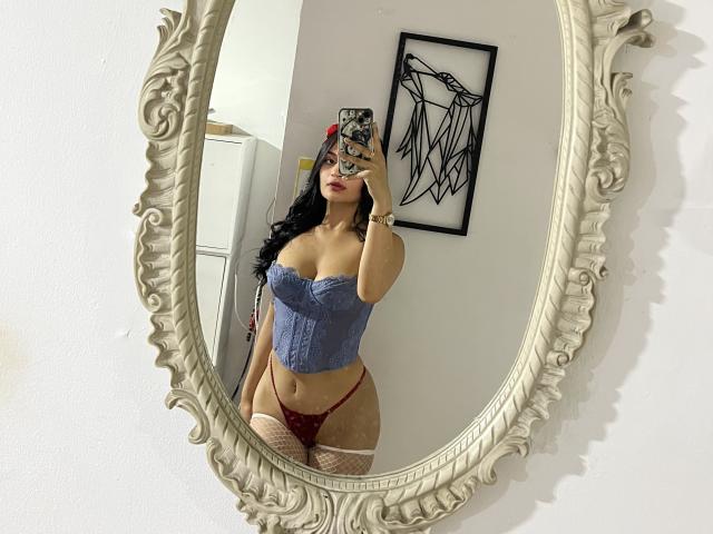 LizzyMarhall69 with dark hair, wearing a blue lace-up corset, red thong, and fishnet stockings, taking a mirror selfie, showing her tits and pussy in the reflection.