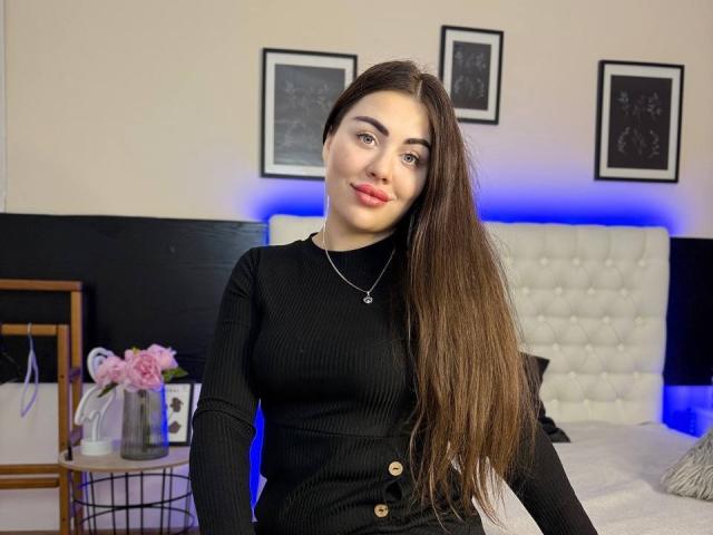 GrayLunna, con cabello castaño largo, vestida con un vestido negro de textura acanalada, sonriendo, con lápiz labial rosa, un collar, y un dormitorio azul con cabecera blanca y rosas rosadas sobre una mesa..