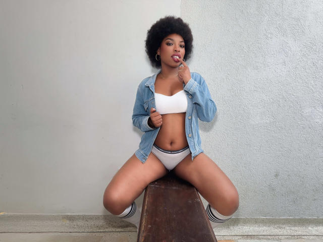 NatashaSparks with a big afro, white bra, denim jacket, and white panties, sitting on a wooden stool, sucking a lollipop, legs spread, wearing knee socks.