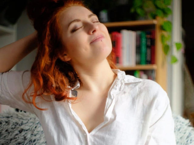 RubyLust-hot with red hair, eyes closed, smiling, wearing a white button-up shirt, head tilted, hand in hair, sitting on a fluffy gray surface, looking relaxed and happy.