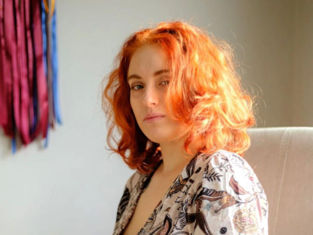 RubyLust-hot with red curly hair, wearing a floral top, has a subtle, confident expression. Her tits are small, and she's sitting in a sunlit room.