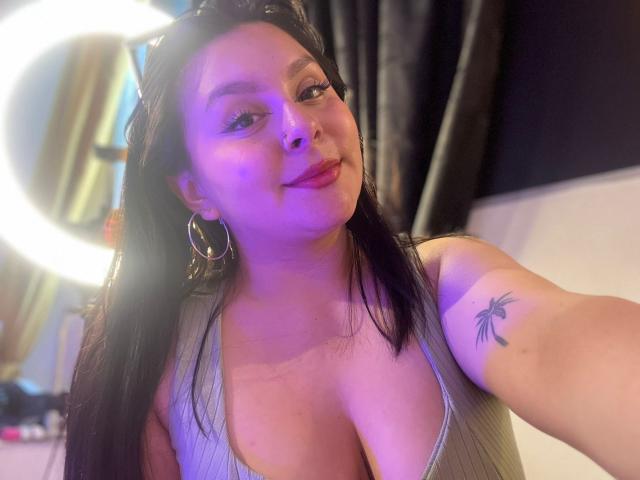 EmaJames with long black hair, purple lighting, large tits in a grey ribbed top, hoop earrings, smiling, and a palm tree tattoo on her right arm.