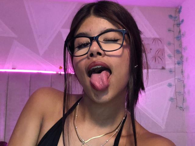 SylvyBrown met een bril, tong uit, ogen gesloten, zwarte haltertop, zilveren ketting, middelgrote borsten, bruin haar, roze licht, en een blauwe muur met bloemenstickers op de achtergrond..