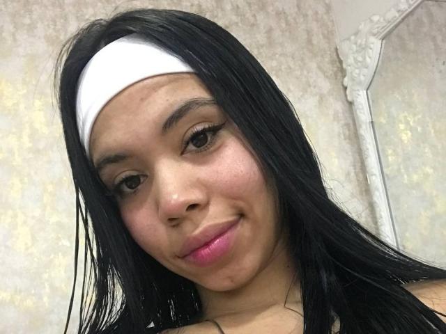 EmaGarces has straight black hair, a white headband, and pink lipstick. Her brown eyes are slightly upturned, and she's smiling subtly. Her ass is not visible.