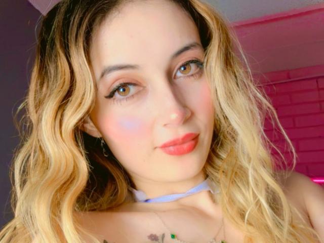 MelissaWendy has blonde, wavy hair, blue eyes, pink lipstick, and a white choker. Her tits are slightly visible with a green tattoo above her cleavage. She's smiling.
