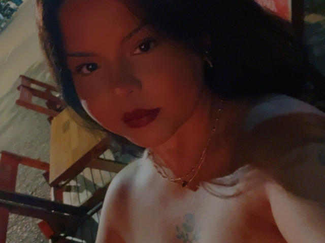 Tryss tiene el pelo oscuro, lápiz labial rojo y lleva un collar dorado. Está sin ropa de arriba, con senos pequeños y un tatuaje en el pecho. La iluminación es tenue, y está mirando a la cámara..