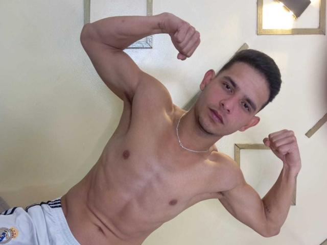 AngelDusstt es un hombre musculoso, sin camisa, con cabello corto y negro, que flexiona ambos bíceps. Lleva pantalones blancos con rayas negras y una cadena de plata..