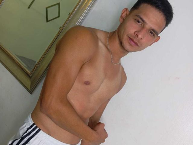 AngelDusstt, sem camisa, com bronzeado, corpo musculoso, cabelo preto curto e um colar de prata, vestindo calças brancas com listras pretas, em pé com um leve sorriso e os braços cruzados..