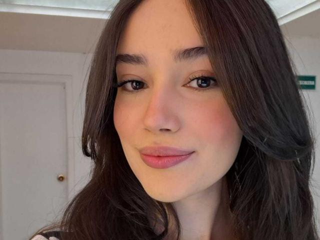 O rosto de MariaLoft, com cabelos castanhos longos, pele clara e lábios rosados, sorrindo suavemente, mostrando uma maquiagem discreta e uma expressão relaxada, em uma imagem no estilo de selfie..