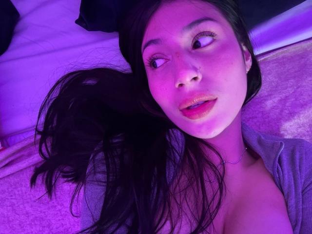 SophieBeaumont's face with purple-lit skin, long black hair, nose ring, and glossy pink lips, wearing a gray shirt, showing a little cleavage, looking to the side.