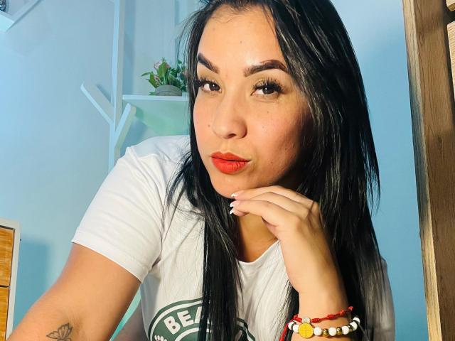 SalomeMilleer with long black hair, red lipstick, and white tee, pouting slightly, hand on chin, wearing a red and white bracelet, has a tattoo on her right arm.