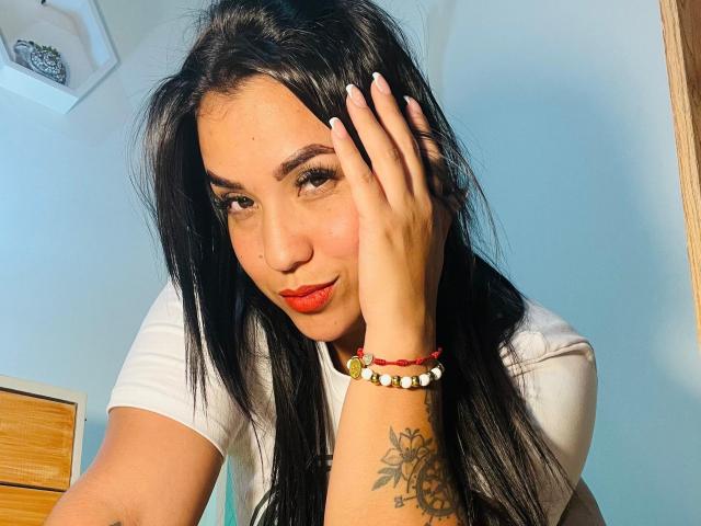 SalomeMilleer with black hair, red lipstick, white shirt, hand on head, wrist bracelet, tattoo on forearm, looking at camera, slightly smiling, showing off her cute face and tits.