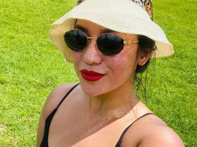 SalomeMilleer with a leopard-print hat, black sunglasses, red lipstick, and black strappy top, standing on green grass, showing her cute face and small tits.