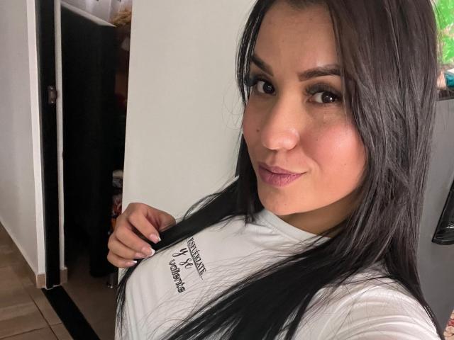 SalomeMilleer has long black hair, brown eyes, and is wearing a white shirt, showing off her tits with a slight smile and purple lipstick. She's holding her hair.