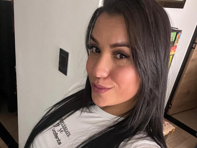 SalomeMilleer has long black hair, brown eyes, and a slight smile. She's wearing a white shirt, showing her tits. Her skin is tan and she has pink lipstick.