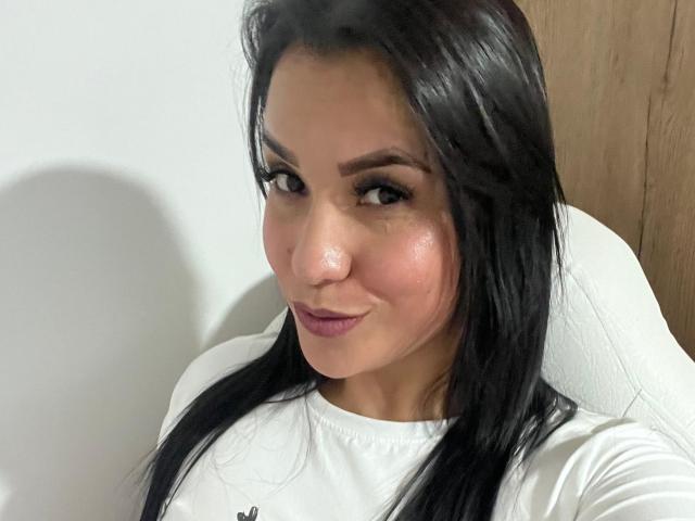 SalomeMilleer has shiny black hair, a slightly flushed face, and a subtle smile. She's wearing a white shirt, sitting with a wooden door behind her.