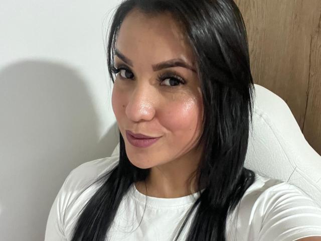 SalomeMilleer has shiny, slightly flushed skin, black hair, and is wearing a white shirt, with a subtle smile, showing her tits. She's looking at the camera.
