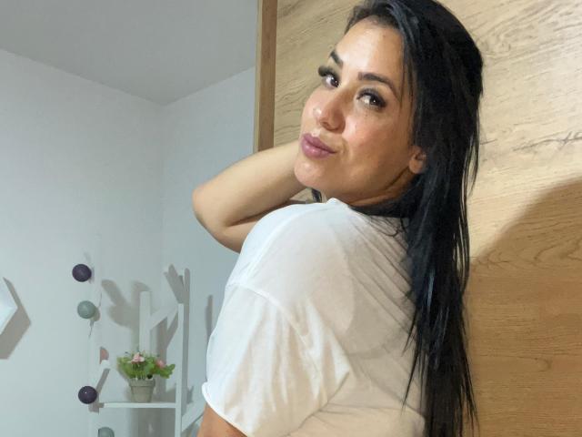 SalomeMilleer with long black hair, white shirt, and pink lipstick, looking over shoulder, hand in hair, smiling slightly, standing against wooden door. Her tits are partially visible.