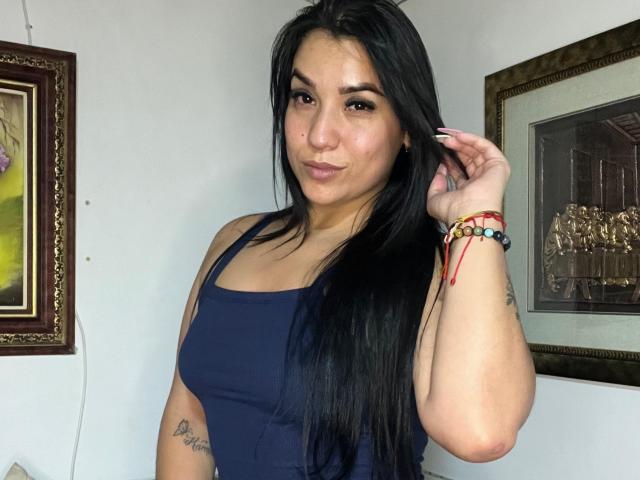 SalomeMilleer with long black hair, dark eyes, and small tits, wearing a navy tank top, smiling slightly, touching her hair, and wearing colorful bracelets, in a photograph.