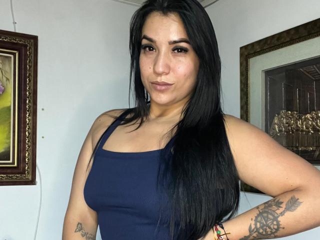SalomeMilleer with long black hair, wearing a blue tank top, showing her tits, a tattoo on her right arm, and a colorful bracelet, looking at the camera.