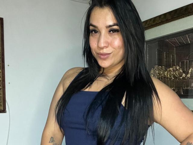 SalomeMilleer has long black hair, a small tattoo on her right arm, and is wearing a dark blue tank top, showing her tits. She's smiling at the camera.