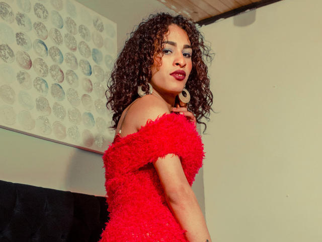 StefannyMia in a red, off-shoulder, textured dress with curly brown hair and red lipstick, wearing large hoop earrings, looking over her shoulder with a slight smile.