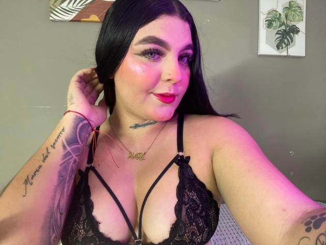 MelodyBlossom con reggiseno in pizzo nero, incarnato rosato, rossetto rosso, tatuaggi su braccio e petto, lunghi capelli neri, mano tra i capelli, illuminazione viola-rosa, indossa una collana, sorridente..