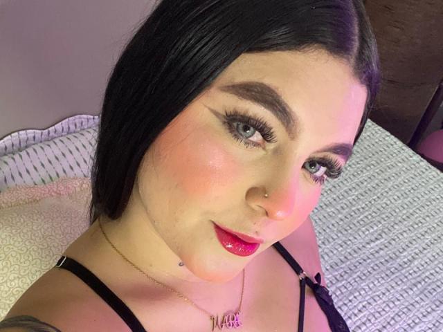 MelodyBlossom has black hair, green eyes, and pink blush. She wears red lipstick, black strap top, nose ring, and necklace. Her tits are small, and she has a cute face.
