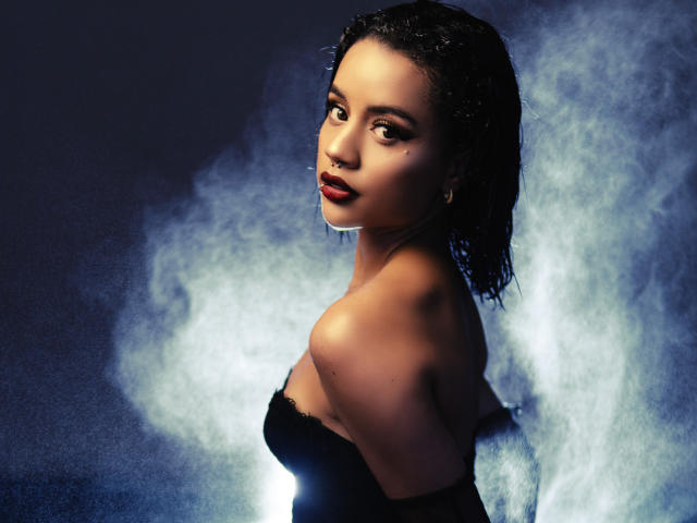 Wendy James, avec ses cheveux noirs et mouillés, sa robe noire sans bretelles et son rouge à lèvres rouge, regardant par-dessus son épaule, une fumée bleu clair et blanche en arrière-plan, une expression sérieuse, plan moyen, sexy..