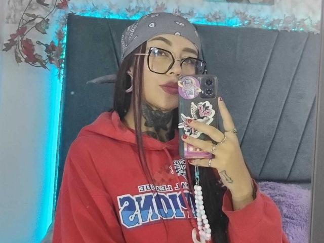 Amber Keller, com cabelos castanhos longos, óculos, moletom vermelho, bandana cinza, tatuagens, segurando um celular com adesivos, unhas roxas, um anel e uma pulseira de contas, em frente a um espelho..