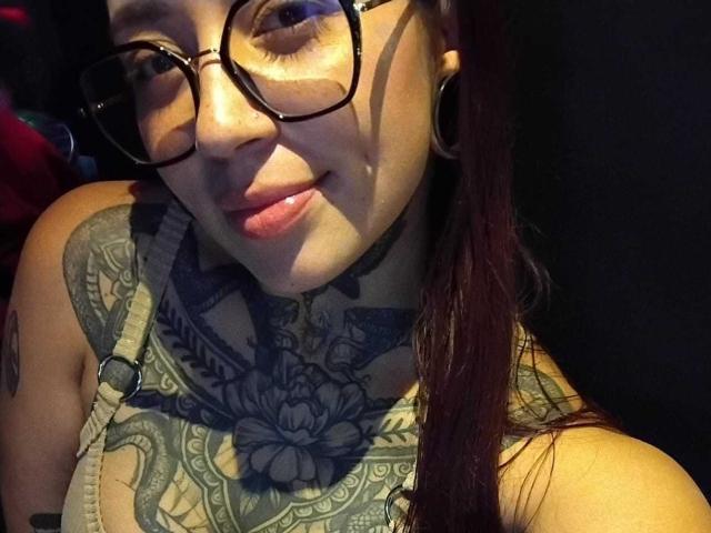 AmberKeller with glasses, dark red hair, and tattoos, smiling at the camera, showing a detailed black and gray chest tattoo, wearing a strap top.