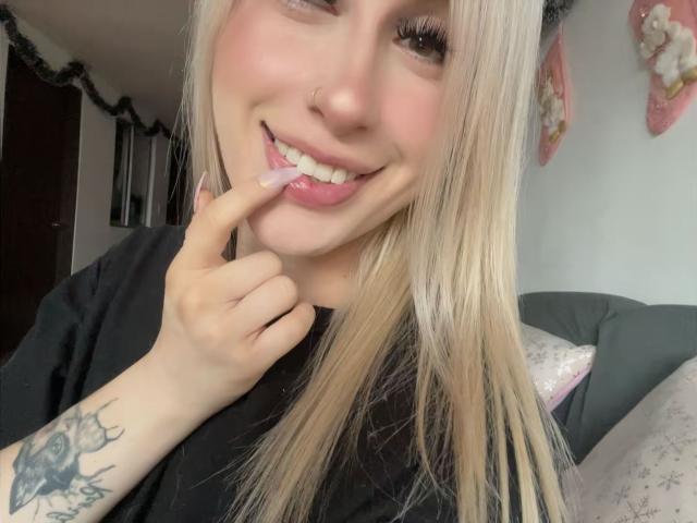 SammyViolet, con cabello rubio, camisa negra, piercing en la nariz y tatuaje en el antebrazo derecho, sonriendo y tocándose los labios rosados con el dedo, mostrando sus dientes blancos..