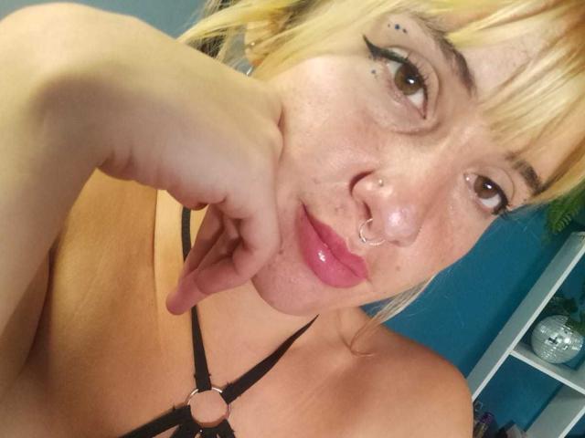 SamanthaaMiller with blonde hair, nose ring, and lip ring, pouting with pink glossy lips, black harness, and hand on cheek, showing freckles and black eyeliner.