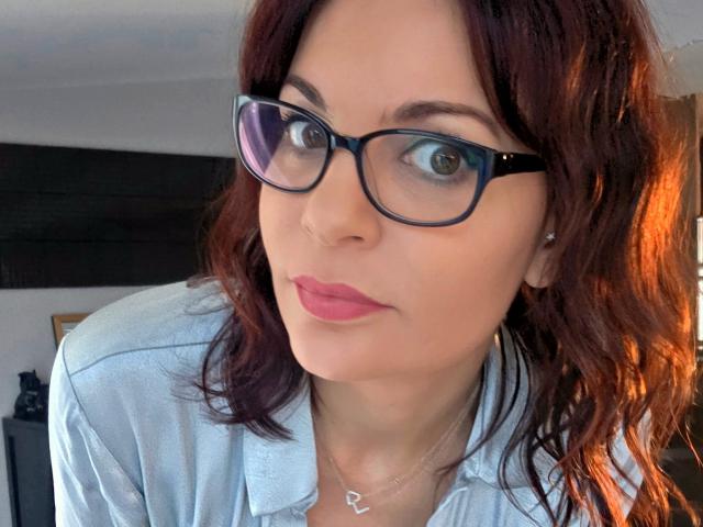 Nahaufnahme von JuneLaFrenchie, einer Frau mit hellbrauner Haut und schulterlangem, braunem Haar, die eine schwarze Brille und ein weißes hemd mit Knopfleiste. Sie blickt direkt in die Kamera und lächelt dabei leicht. Verdammt heiß..