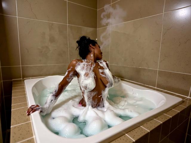 Cocohot-hot is in a white bathtub with bubbles, naked, dark skin, soapy tits, and back, sitting with arm on edge, steam rising, hair in a bun, tiled walls.