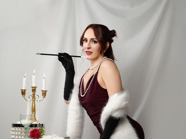ButtyCherry, con cabello rojo recogido en un moño, vestida con un vestido rojo oscuro, guantes negros y piel sintética blanca, sosteniendo un cigarrillo, perlas y un candelabro dorado con velas encendidas..