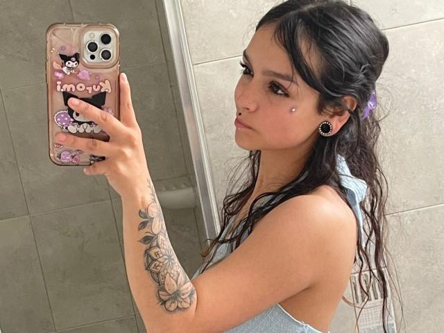 MarietteJones che si scatta un selfie allo specchio, mostrando il seno in una canottiera grigia, con lunghi capelli neri bagnati, una molletta viola per capelli, un braccio tatuato e degli adorabili auricolari neri..