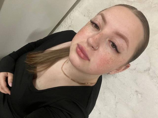 GirlPlayer-hot with freckled face, pink lips, nose ring, and slicked-back brown hair, wearing a black top, gold chain, and subtle eyeliner, in a selfie.