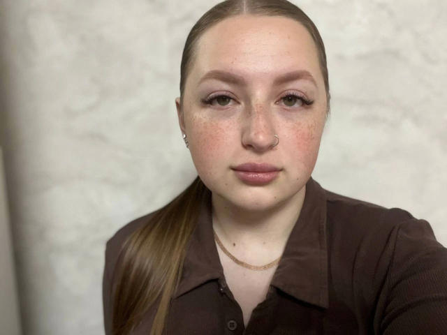 GirlPlayer-hot has light skin, brown hair in a ponytail, brown eyes, a nose ring, and is wearing a brown shirt. Her freckles and subtle makeup are visible.