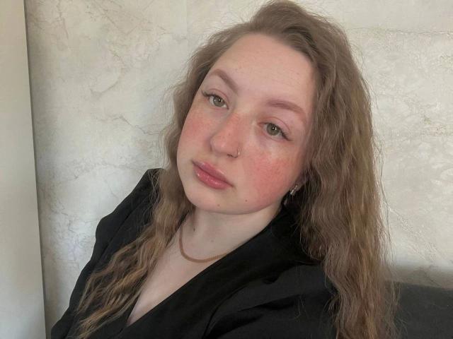 GirlPlayer-hot with wavy brown hair, green eyes, nose ring, and pink freckles, wearing a black shirt, looking slightly to the side with a neutral expression.