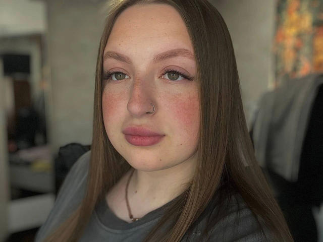 GirlPlayer-hot with straight brown hair, green eyes, pink lips, nose ring, freckles, and gray shirt, looking slightly to the side with a soft, neutral expression.