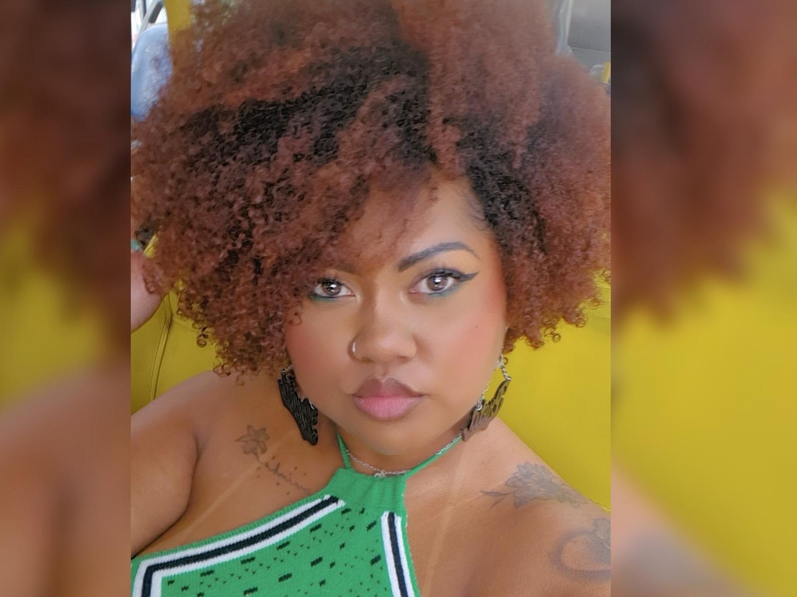 BaiaNinhah with curly brown hair, green halter top, nose ring, tattoos on shoulders, and full lips, looking into mirror, yellow background. Boobs partially visible, black earrings.