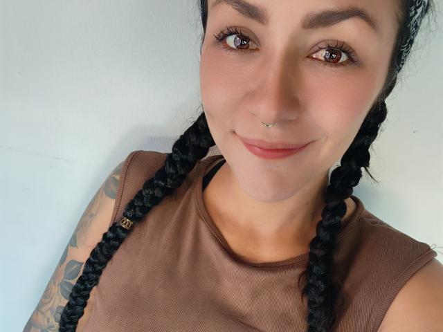 AlejadArte with braided black hair, brown sleeveless top, nose ring, tattooed arm, smiling, brown eyes, light skin, and a subtle pink lip. She's looking at the camera.
