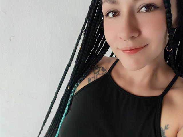 AlejadArte with long black braids, nose ring, and hoop earrings, wearing a black halter top, showing small tits and tattoos on her right arm, smiling softly.