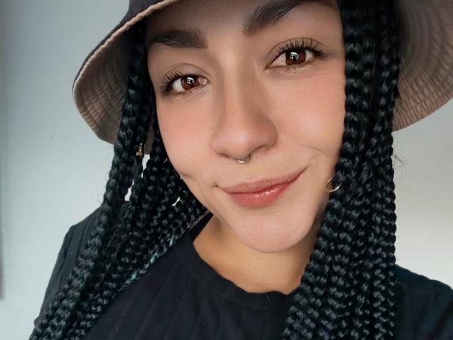 AlejadArte has dark braided hair, brown eyes, a nose ring, and a subtle smile, wearing a beige bucket hat and black shirt, with a cute, attractive face.