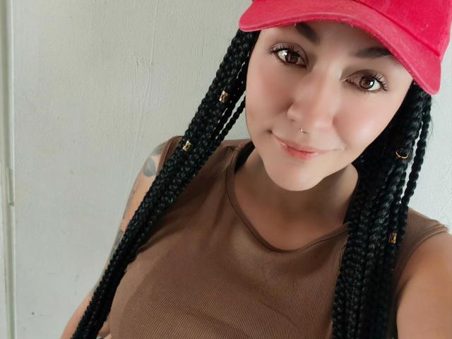 AlejadArte with long black braids, red cap, brown tank top, nose ring, and tattoo on left arm, smiling at camera, showing small, round tits and cute face.