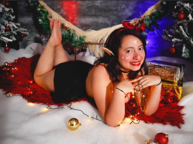 CristinaSmit with elf ears, lying on a red fuzzy rug, black dress, red hairband, smiling, white face paint, red lipstick, black stockings, fairy lights, Christmas ornaments, snow.