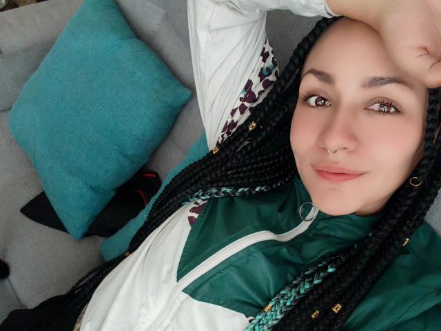 AlejadArte with braided black hair, green and white jacket, nose ring, and cute smile, laying on a gray couch, teal pillow in background, right arm above head.