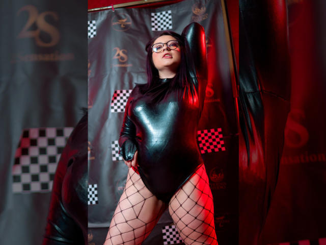 CristinaSmit in a shiny black latex bodysuit, fishnet stockings, and glasses, posing with arm above head, showing her ass and pussy under the red and black lighting.